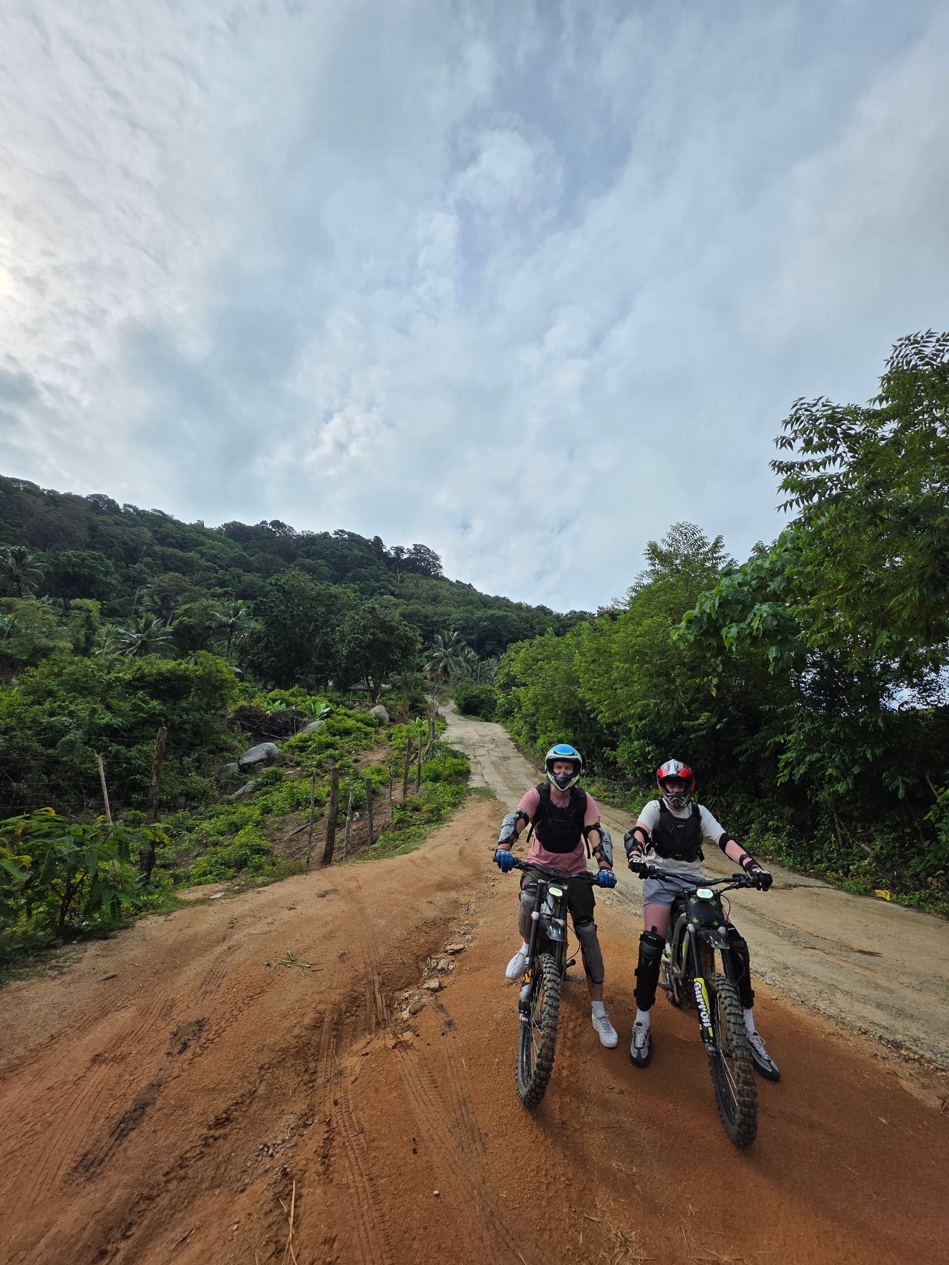 Two individuals riding electric motorbikes on a dirt path surrounded by lush greenery in Phuket, Thailand.
