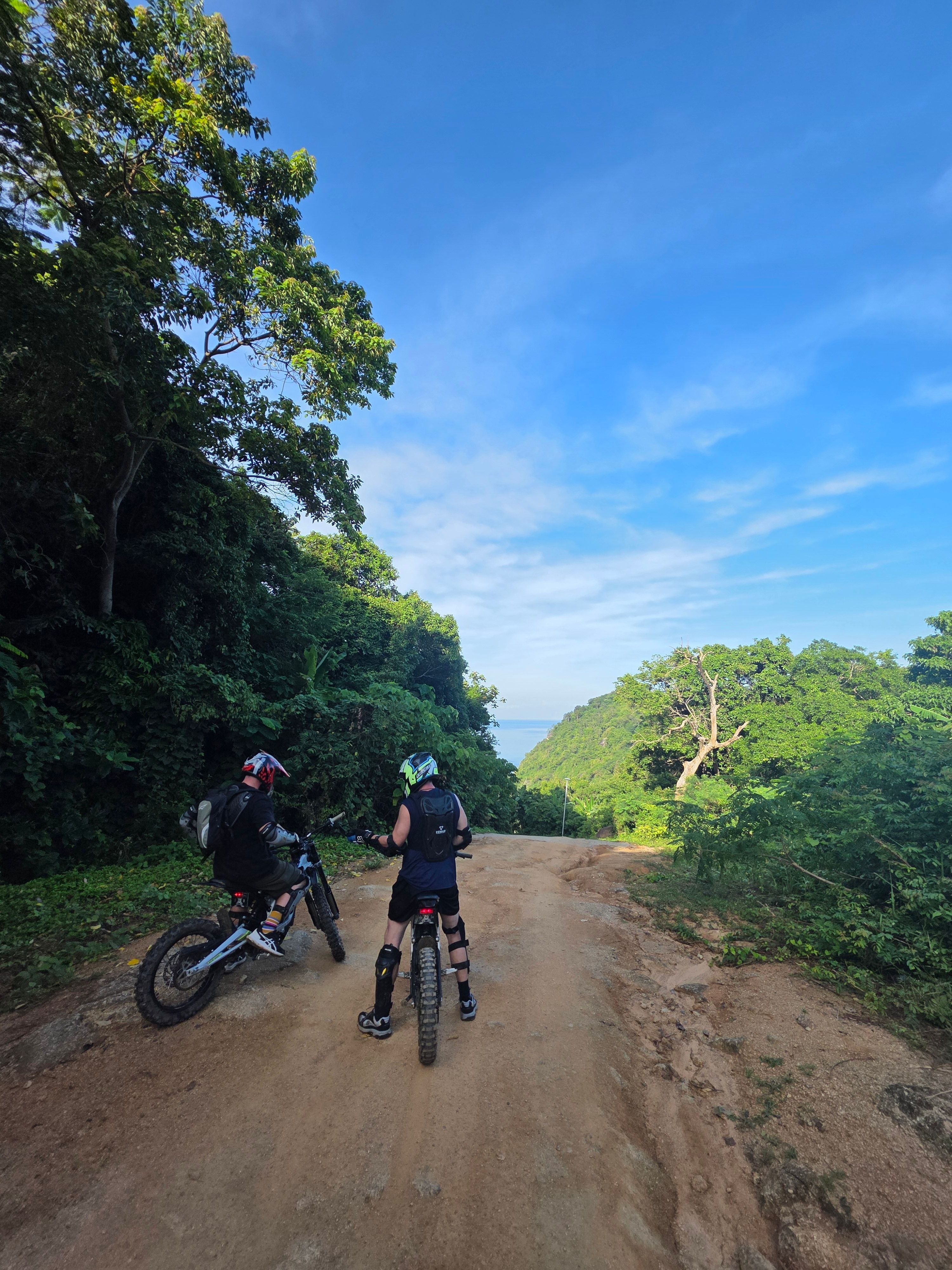 Two individuals riding electric motorbikes on a dirt trail surrounded by lush greenery in Phuket, Thailand.